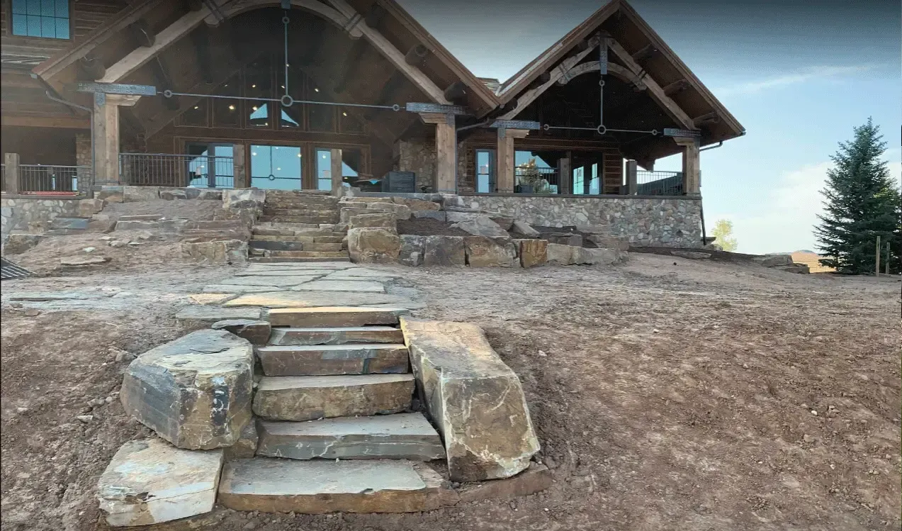 Stone steps leading to a rustic cabin with a wooden porch and steep roof on a dirt hillside.