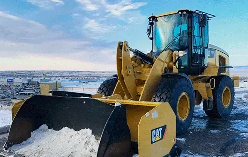 Yellow CAT front-end loader with snow in its bucket, parked on a snowy road under a cloudy sky.