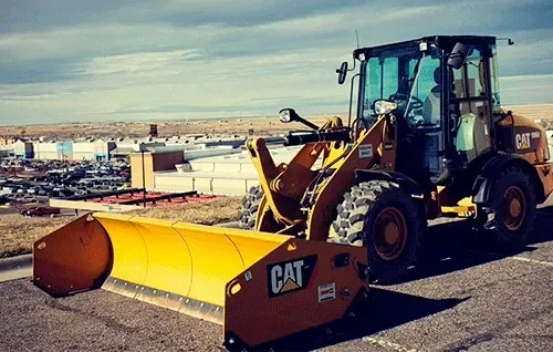 Yellow CAT front-end loader with snowplow attachment parked in a snowy industrial yard.