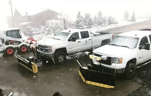 Snow-covered pickup trucks and a plow in a parking lot during heavy snowfall.