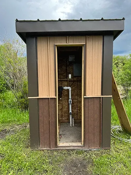 Small wooden outhouse with open door, showing a toilet inside, set in a grassy outdoor area.