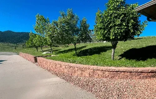 Sunny park path beside red stone wall, green trees, and grassy hillside under a clear blue sky