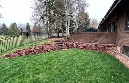 Backyard with green lawn, stacked stone retaining wall, trees, and brick house exterior
