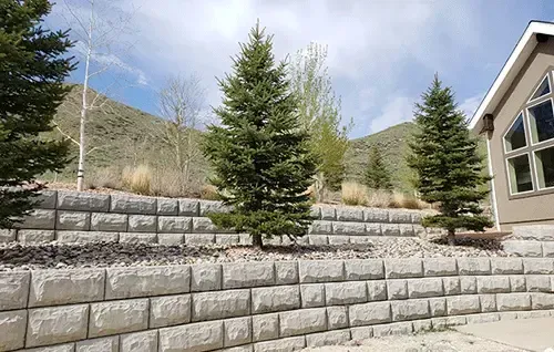 Terraced stone retaining wall with evergreen trees and hills beside a house under a cloudy sky