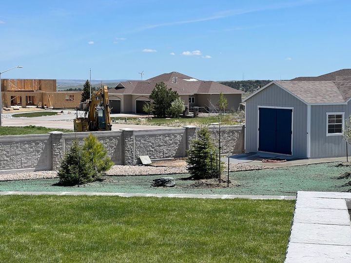 Suburban backyard with green lawn, stone wall, and a gray shed under a blue sky.
