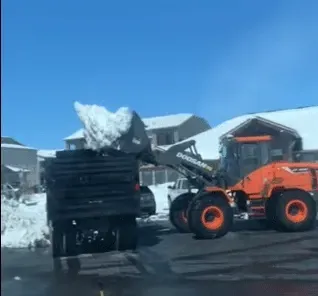 Orange front loader lifting snow beside stacked black pipes on a snowy street with houses in the background