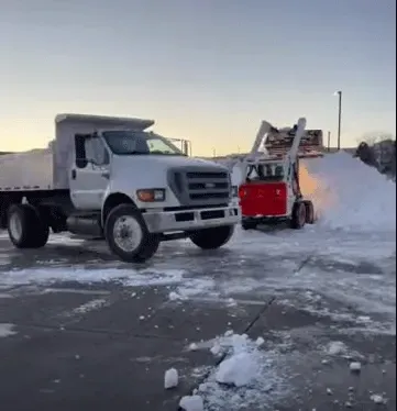 White dump truck and red snowblower clearing snow in a plowed parking lot at dawn