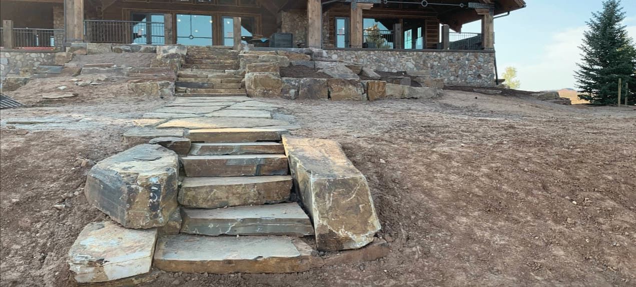Stone steps leading to a rustic lodge on a dirt hillside, with trees and mountains in the background