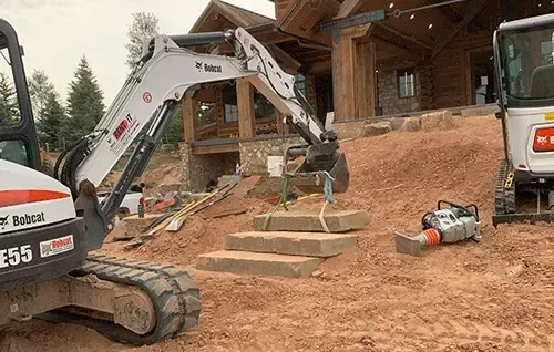 Excavator working beside log cabin steps at a construction site with dirt piles and tools