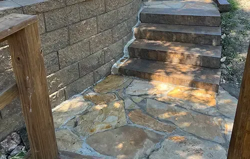 Stone steps beside a retaining wall and wooden handrail in a sunlit outdoor walkway