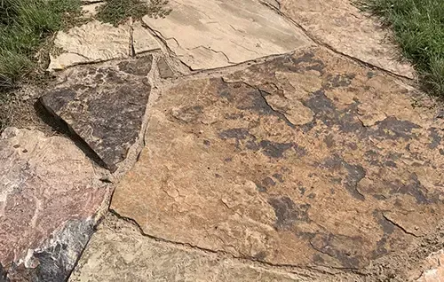Rough stone path with tan and brown slabs outdoors, bordered by grass and dirt