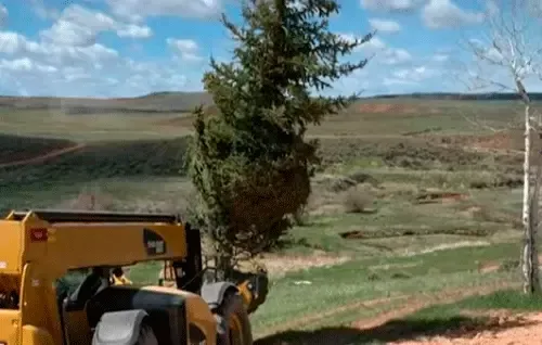 Yellow construction vehicle on a dirt road overlooking a green rural valley with scattered trees and hills.