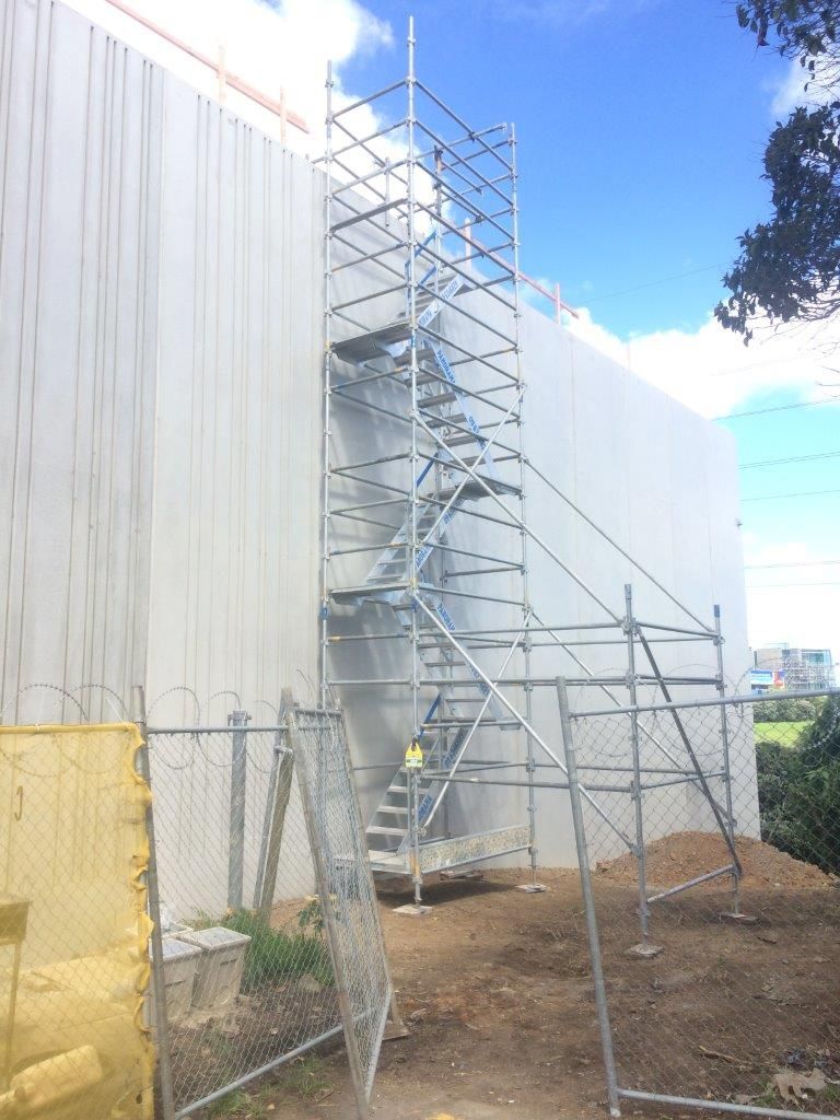 Scaffolding beside a white building wall under a blue sky, with a fenced dirt work area.