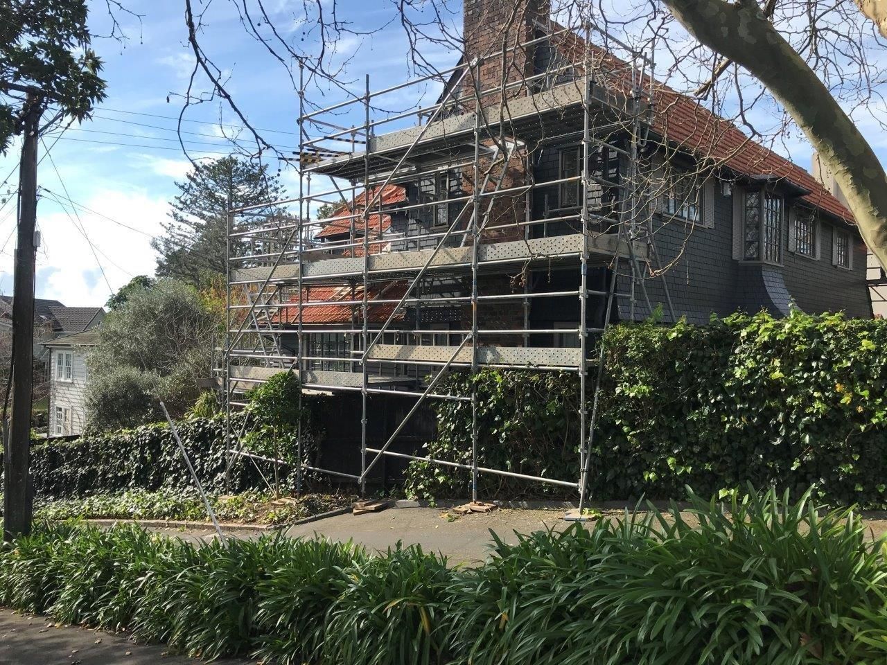 House under renovation with scaffolding, dark siding, and a red-tiled roof surrounded by greenery