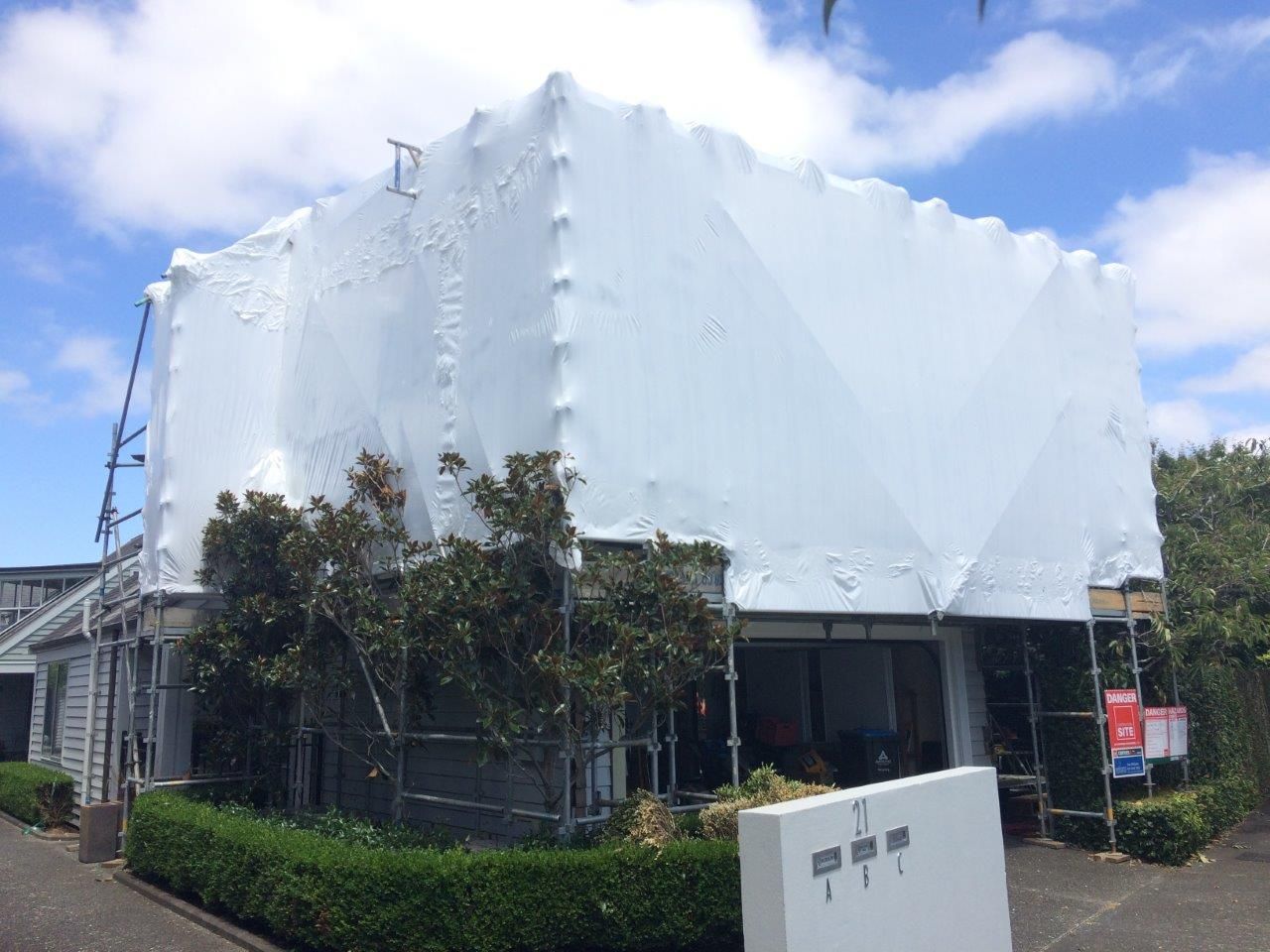 White-wrapped building under renovation beside a hedge and trees on a sunny street corner