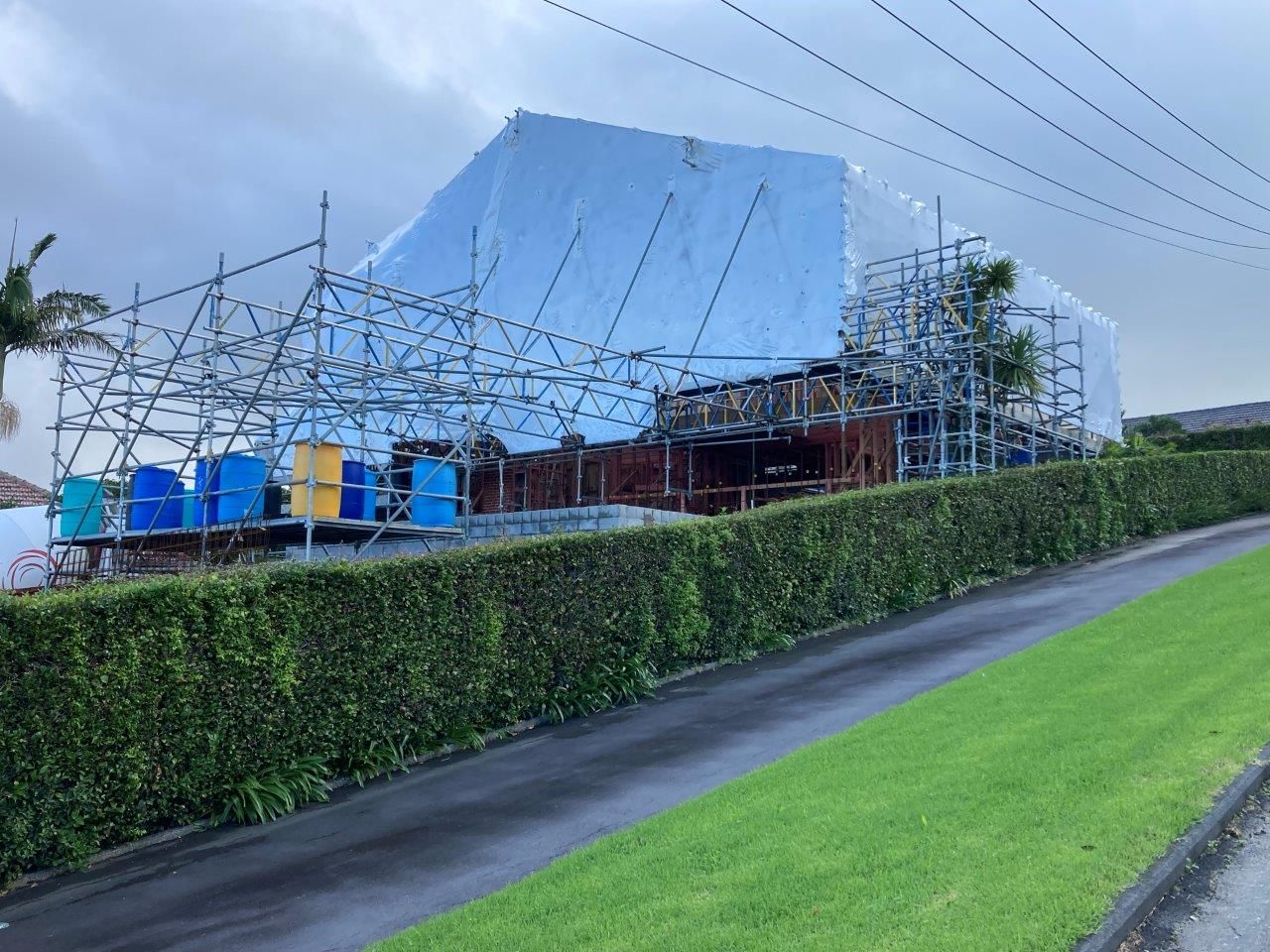 Hedged roadside beside a construction site with scaffolding and blue tarp-covered building materials