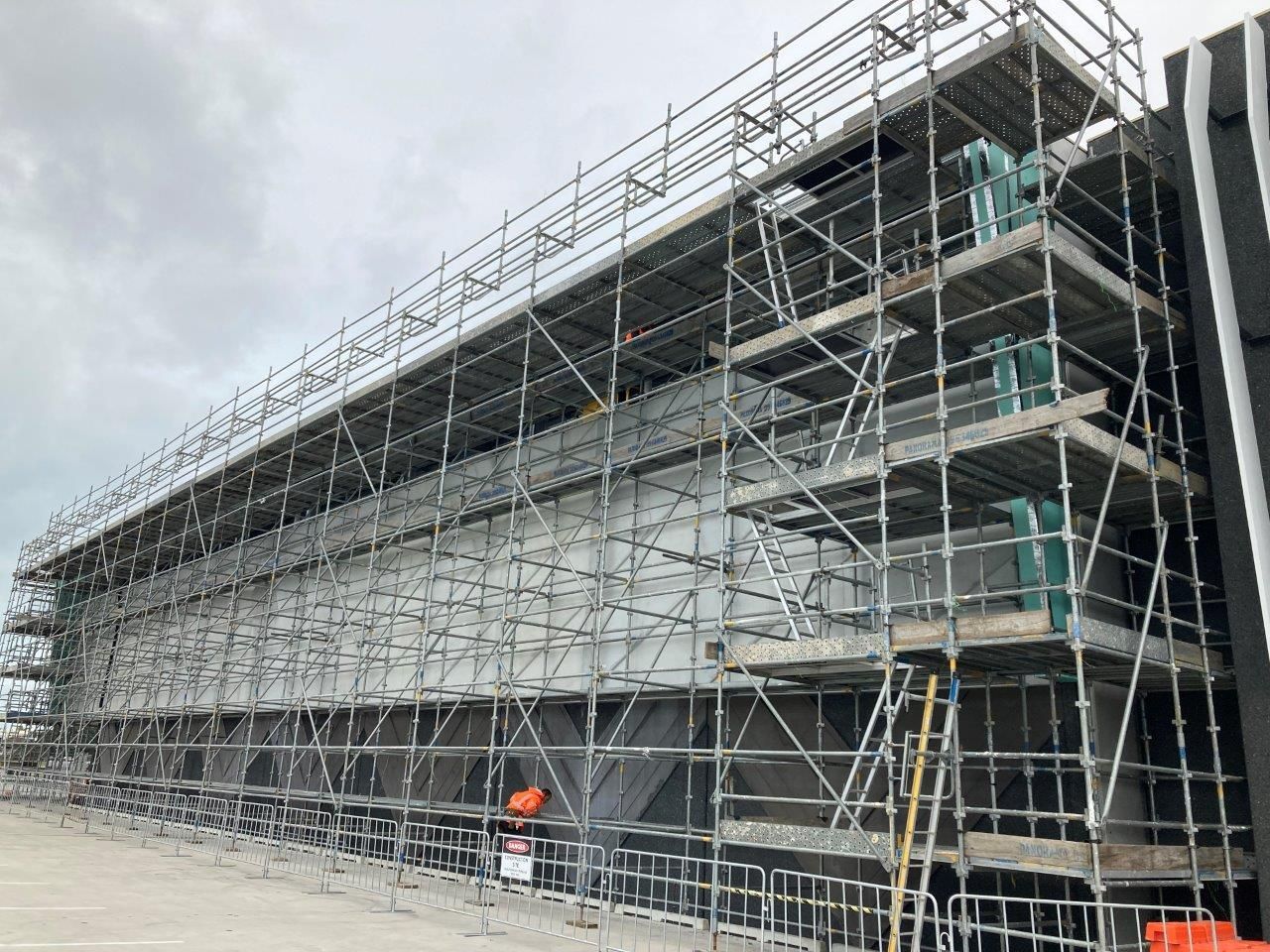 Scaffolding surrounds a large building under construction, with a worker in orange at ground level.