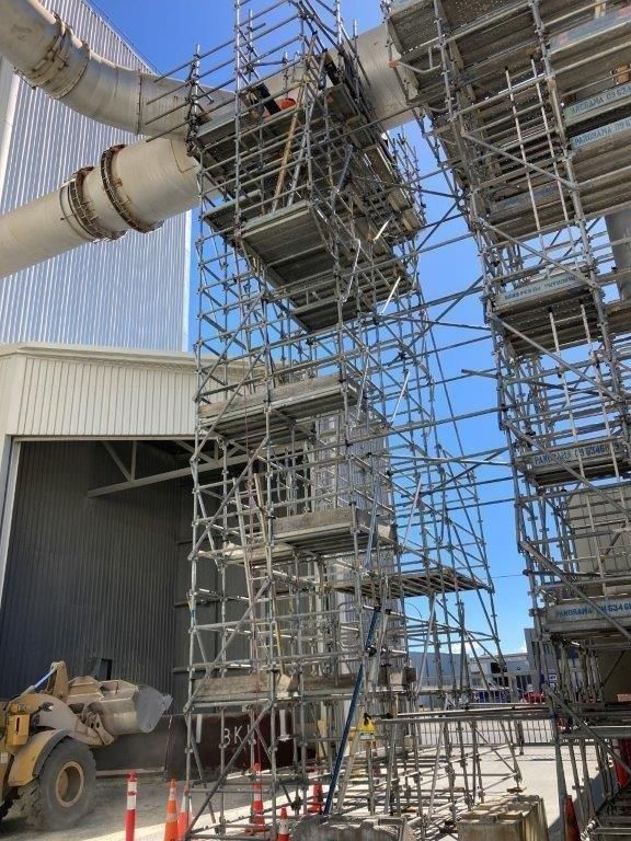 Scaffolding surrounds a large industrial duct beside a building under a clear blue sky.