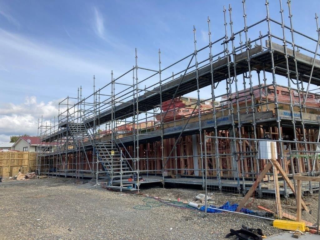 Scaffolding surrounds a row of houses under construction on a gravel lot under a cloudy sky