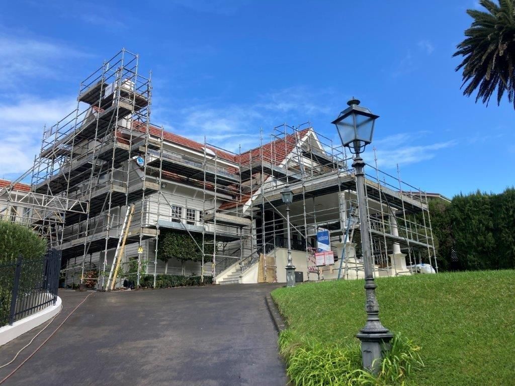 Scaffolding surrounds a large house under renovation beside a grassy lawn and lamppost under a blue sky