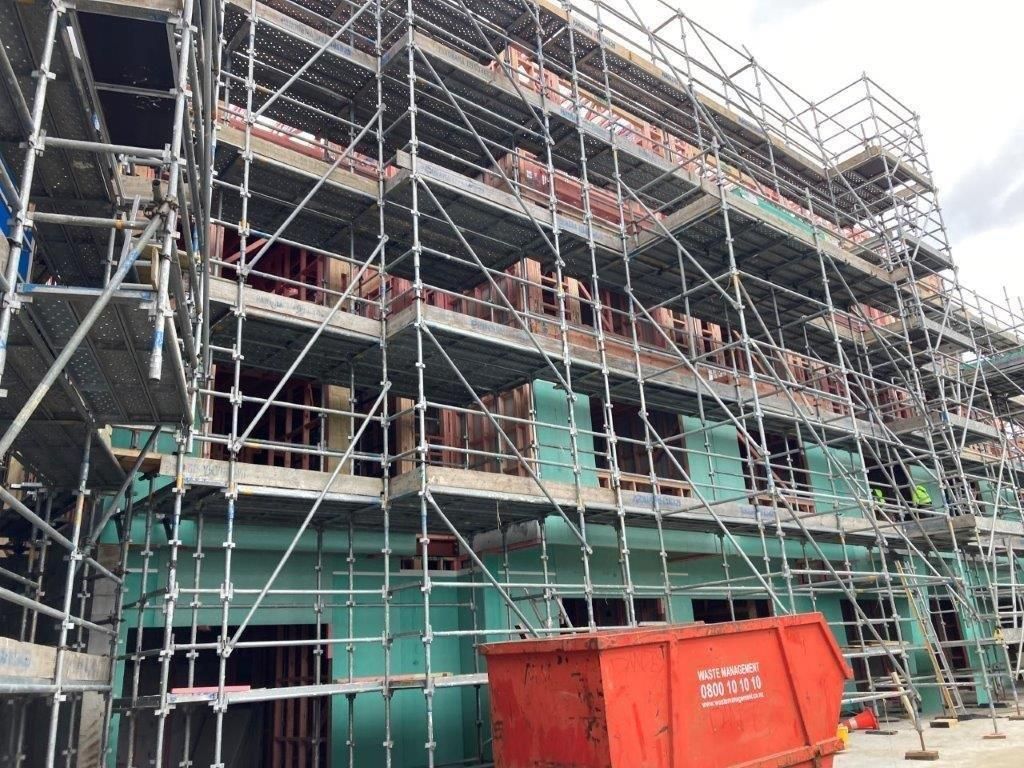 Scaffolding around a building under construction, with green sheeting and an orange dumpster in front