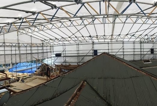 Interior of a large tent structure with exposed trusses and tarps, seen over rooftops and construction materials