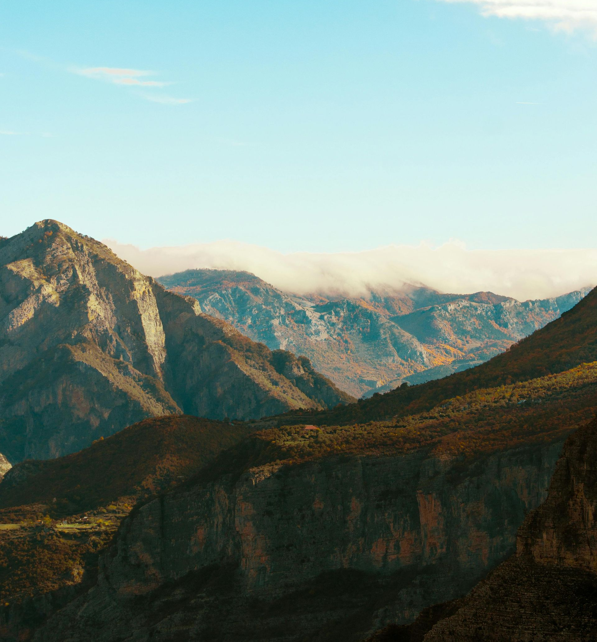 Mountain landscape with sunlit rocky peaks, dark ridges, and low clouds under a pale blue sky