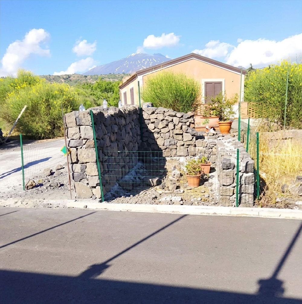 Muro di cinta in pietra con piante in vaso davanti a una casa beige e montagne sullo sfondo, sotto un cielo azzurro.