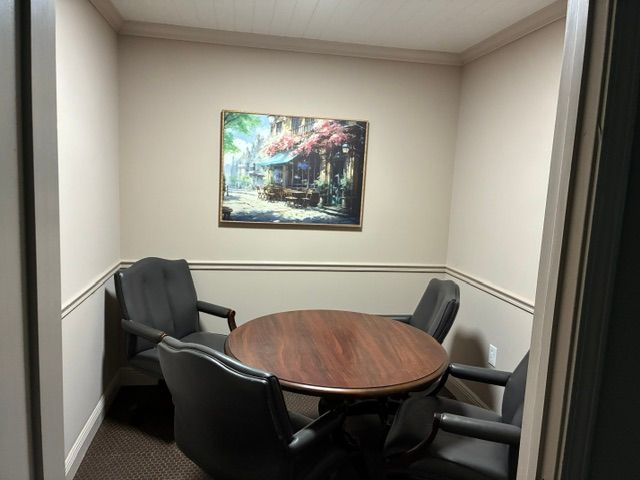Small meeting nook with four black chairs around a round wooden table beneath a framed painting.