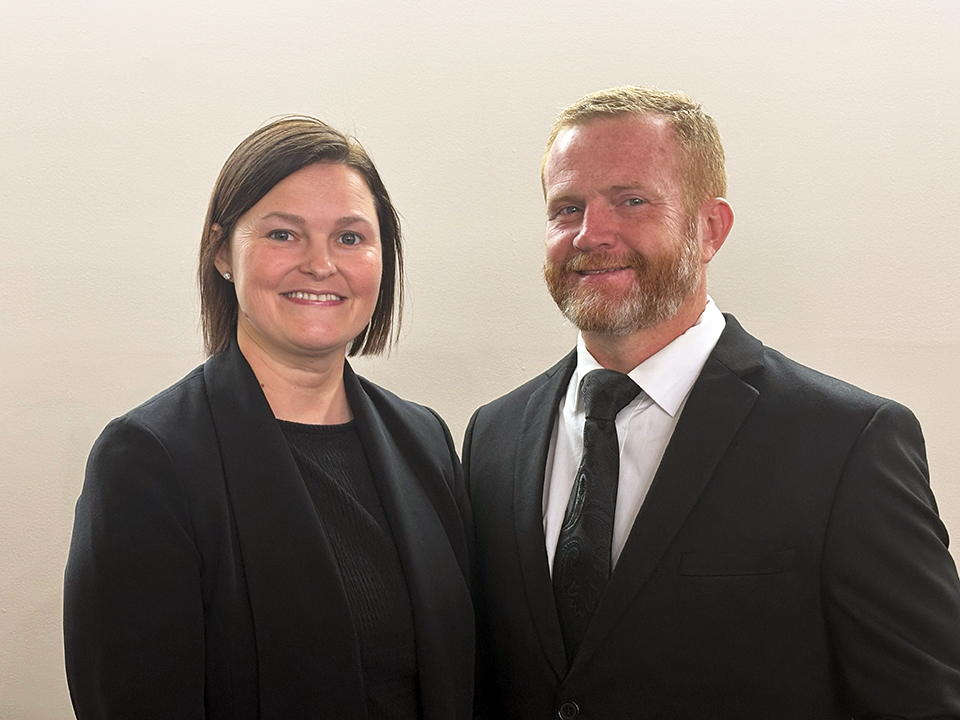 Two people in black suits smiling against a plain light background