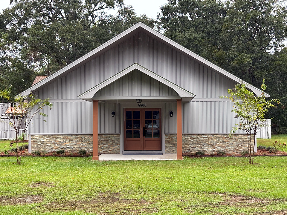 Small gray building with a front porch, stone base, and double wooden doors in a grassy yard.