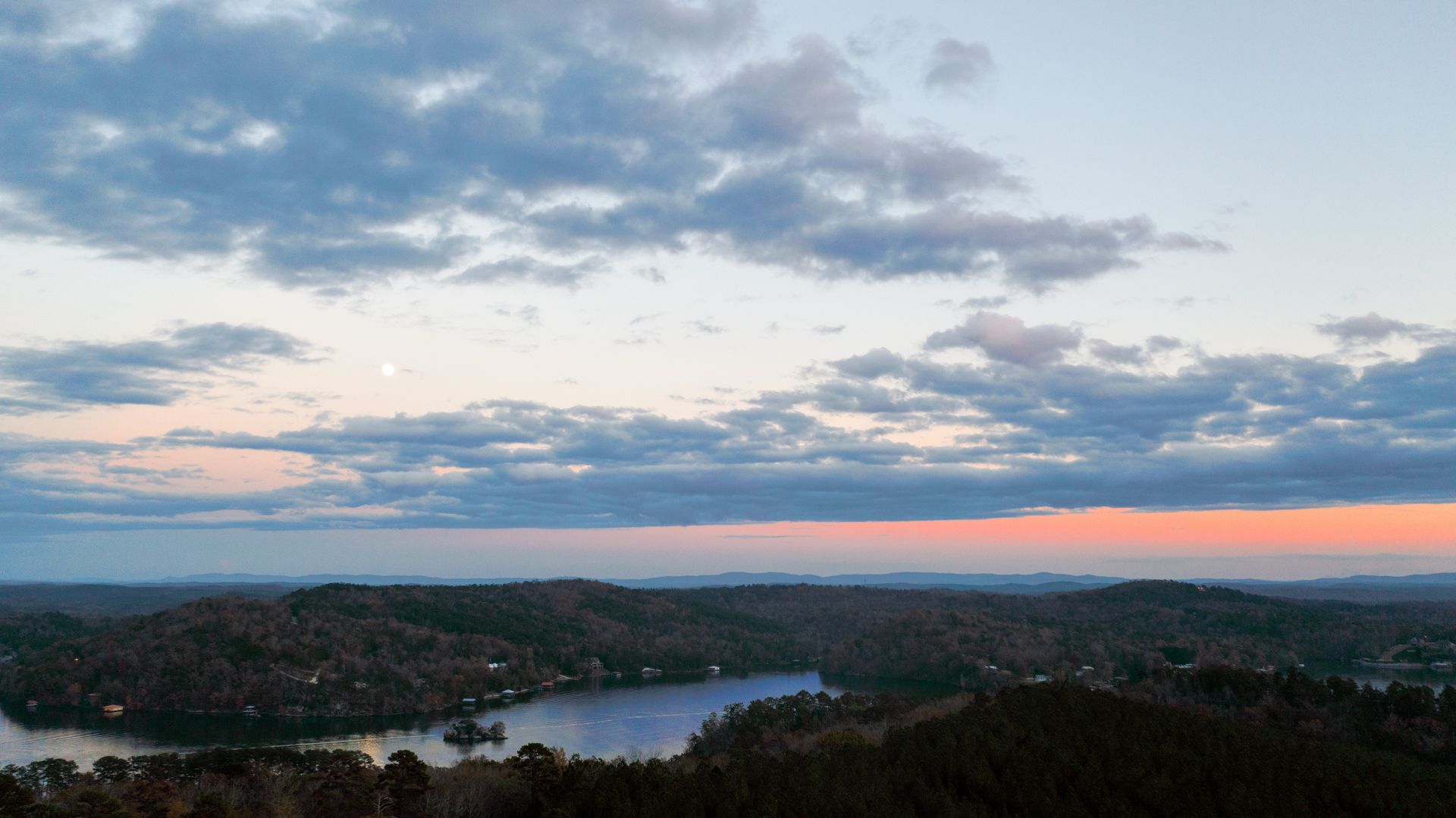 Wide sunset view over a lake and forested hills under a cloudy pink-blue sky