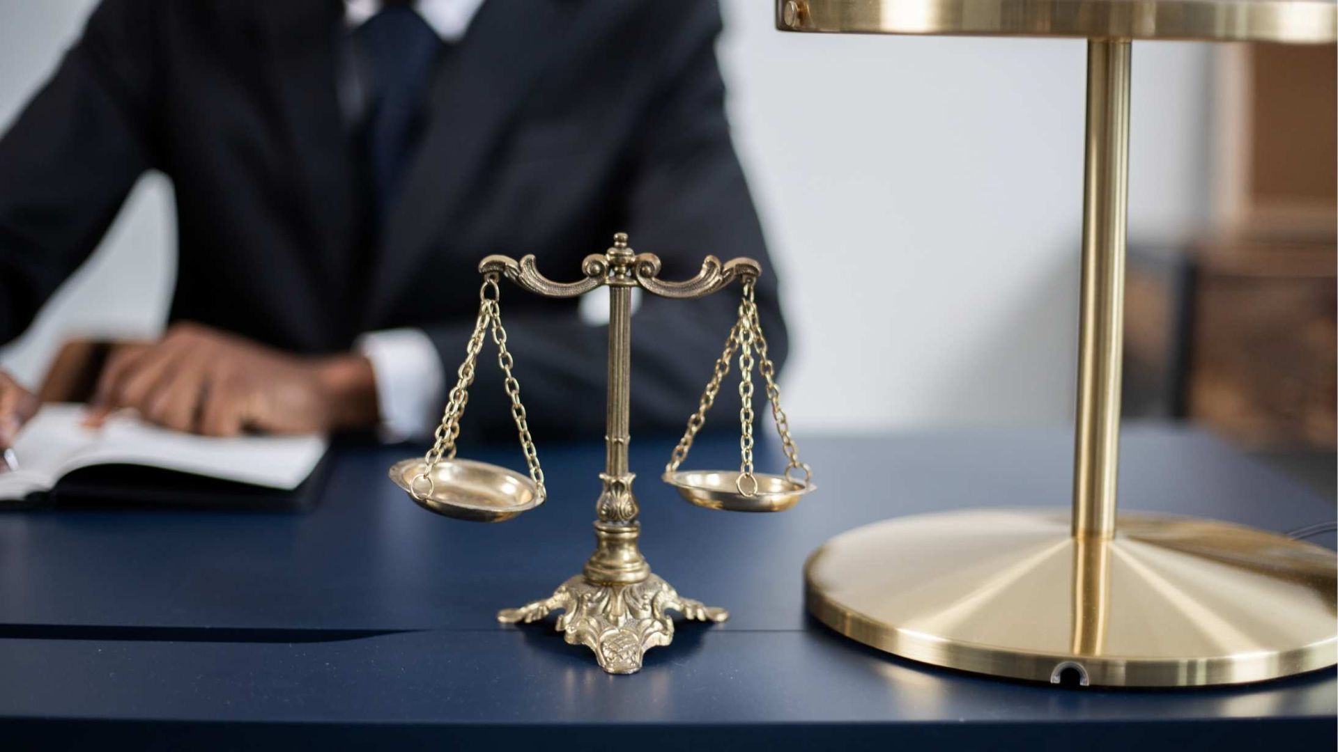 Gold scales of justice beside a brass lamp on a desk with a blurred person in the background