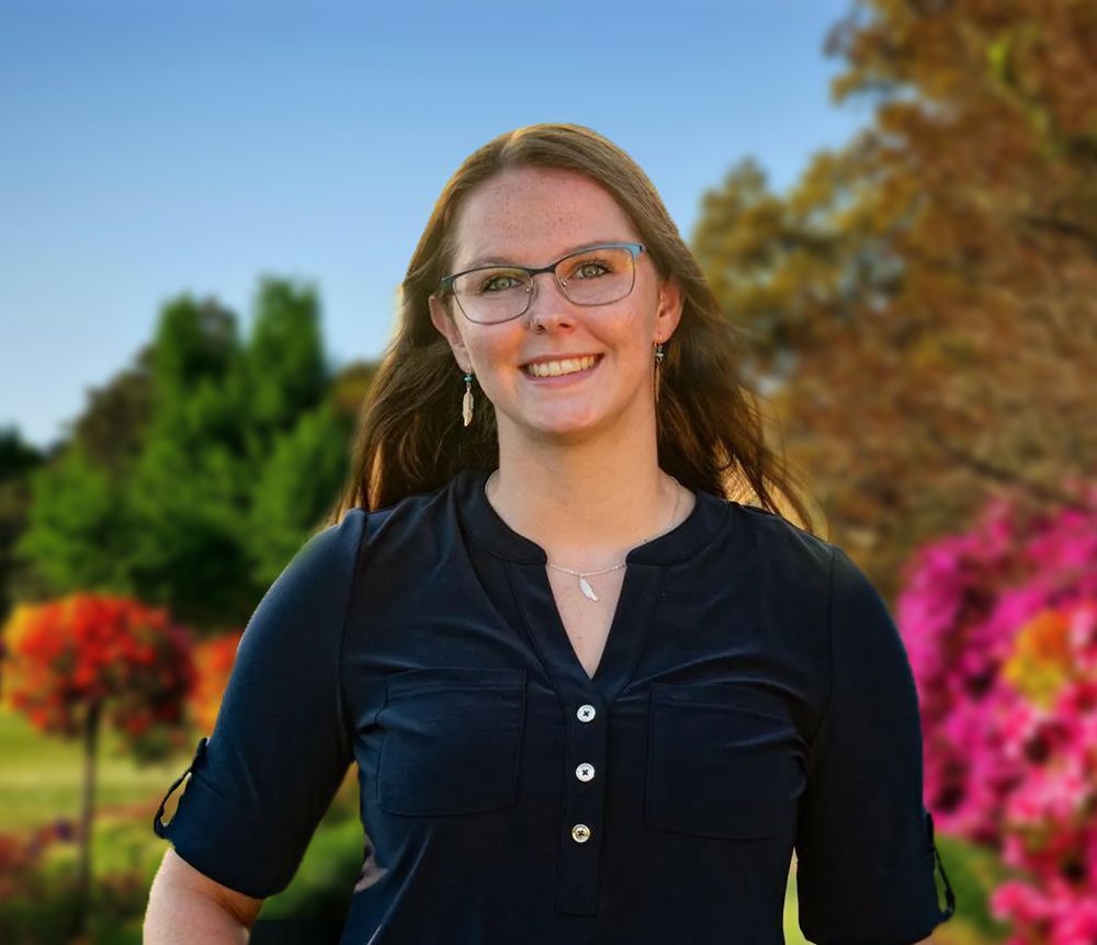 Smiling woman in glasses wearing a dark shirt, standing outdoors with colorful flowers and trees behind her