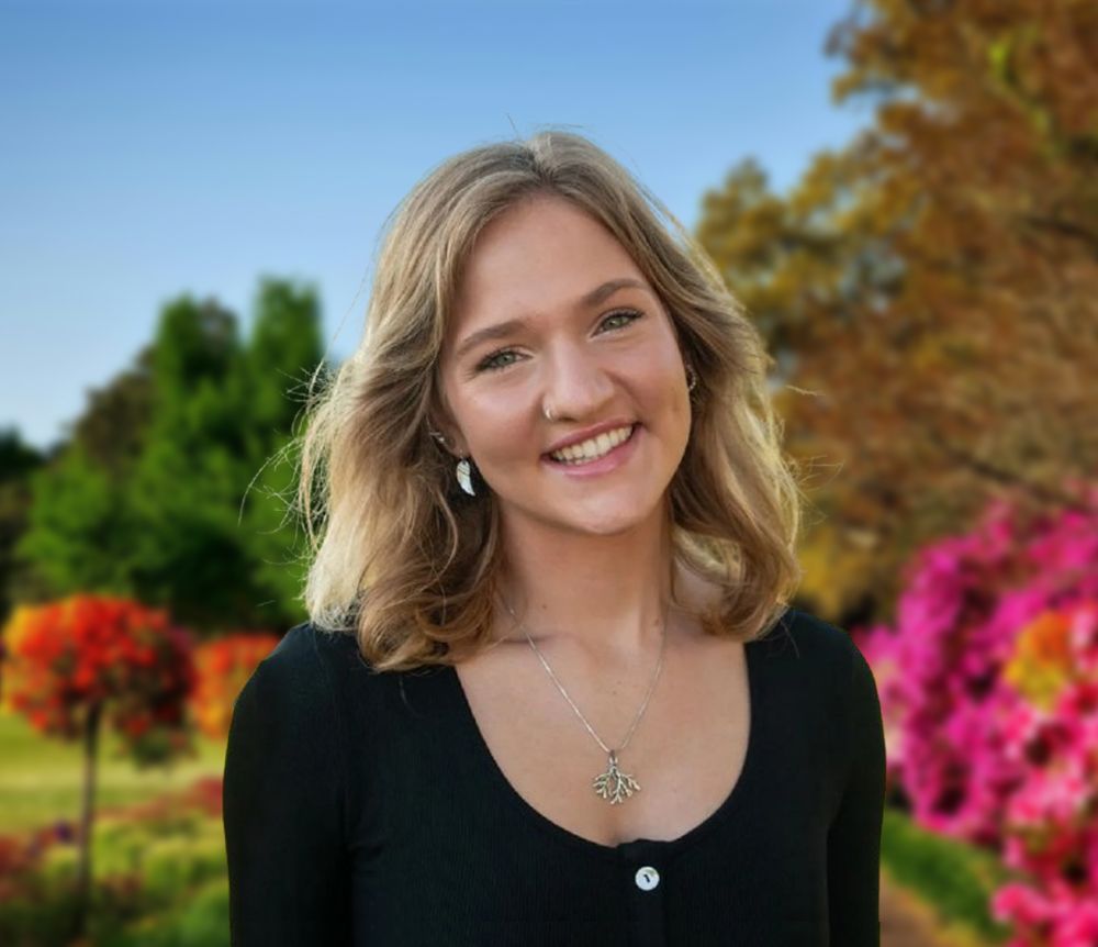 Smiling person in a black top standing in a colorful garden with flowers and autumn trees