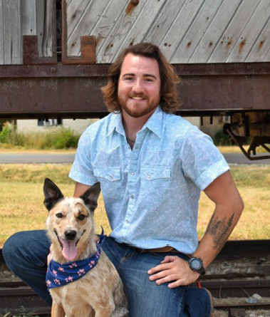 Man kneels beside a tan dog wearing a bandana in front of a wooden railcar.