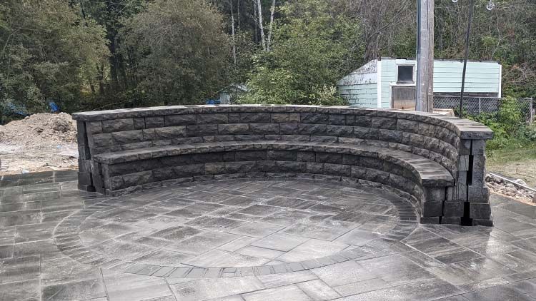 Curved stone bench on a paved patio, with a shed and trees in the background.