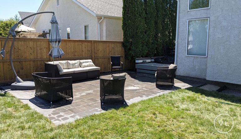 Patio with wicker furniture, umbrella, and a wooden fence. Green grass surrounds paved area.