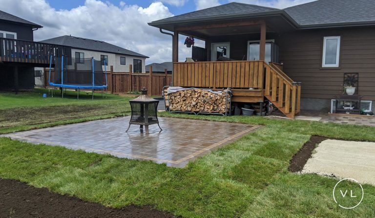 Backyard patio with fire pit, adjacent to a house with wooden deck, surrounded by green grass.