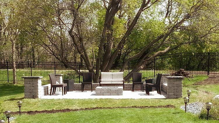Outdoor patio with seating around a fire pit under a tree; surrounded by grass and a black fence.