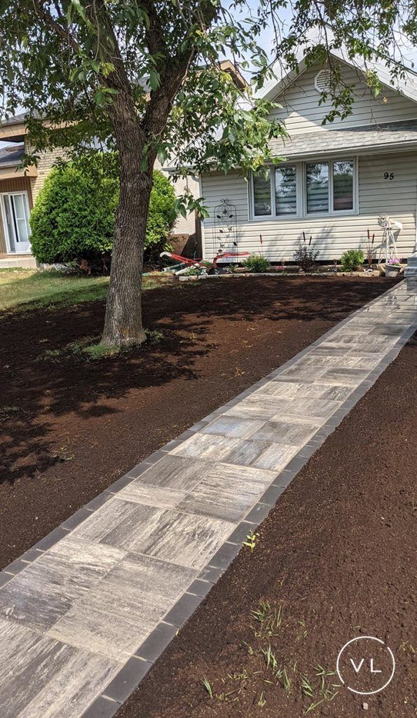 Brick pathway leads to a white house with a tree on the side; brown mulch surrounds the path.