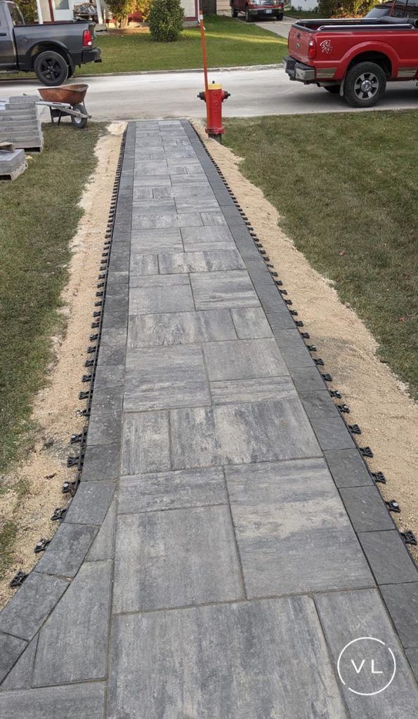 A newly constructed stone paver walkway with black edging, bordered by grass and sand, with a fire hydrant.