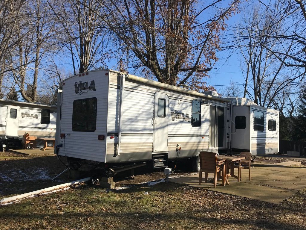 White RV trailer parked on a dirt lot beside a small table under leafless trees and a blue sky
