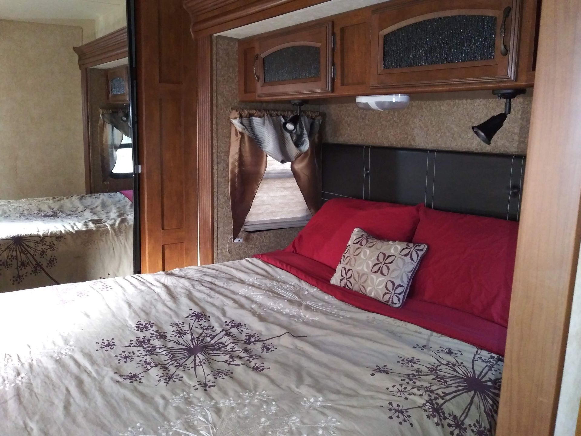 Cozy RV bedroom with a made bed, red pillows, wood cabinetry, and a window curtain.