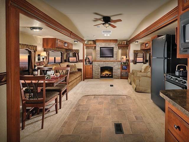 Narrow RV living room with beige seating, wood cabinets, TV, fireplace, and kitchen area.