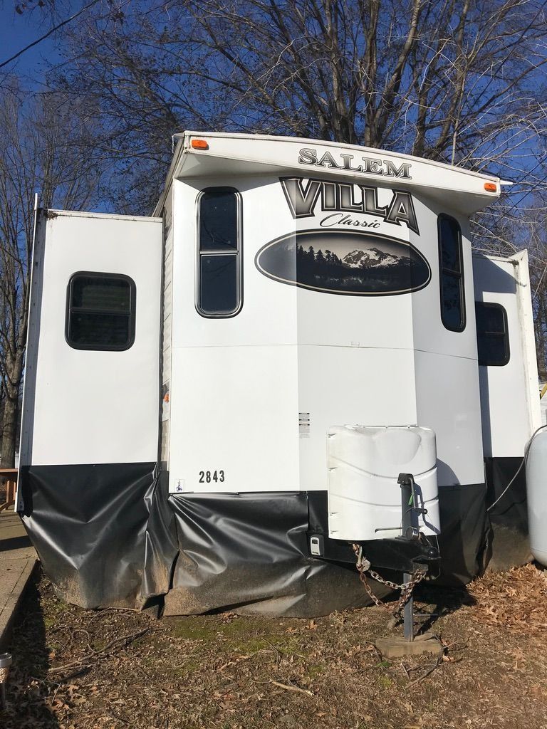 Small white camper trailer with black lower trim parked outdoors among bare trees.