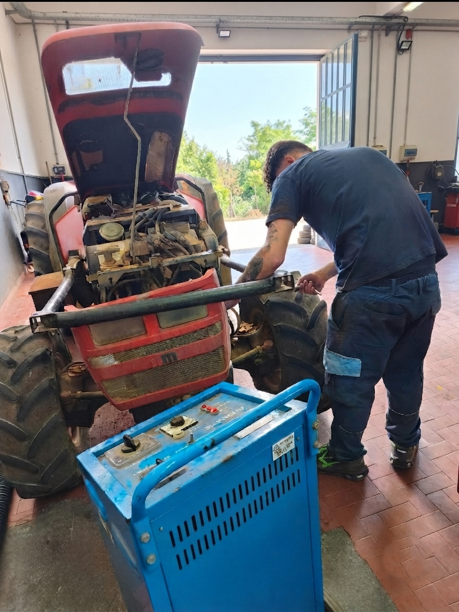 Un uomo lavora al motore di un'auto rossa in un garage, con un macchinario blu in primo piano.