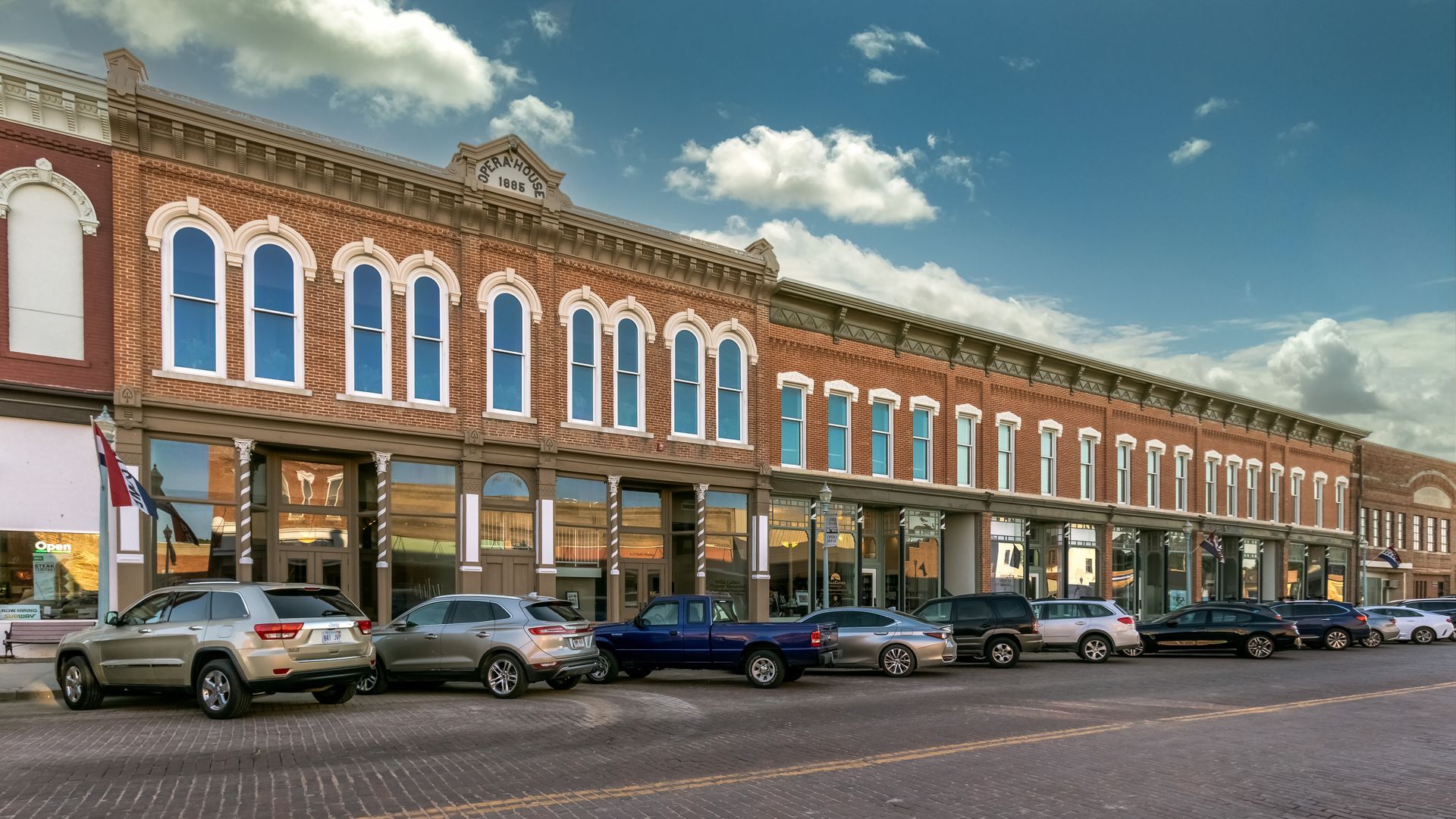 Historic brick storefronts with ornate facades and parked cars along a street under a blue sky
