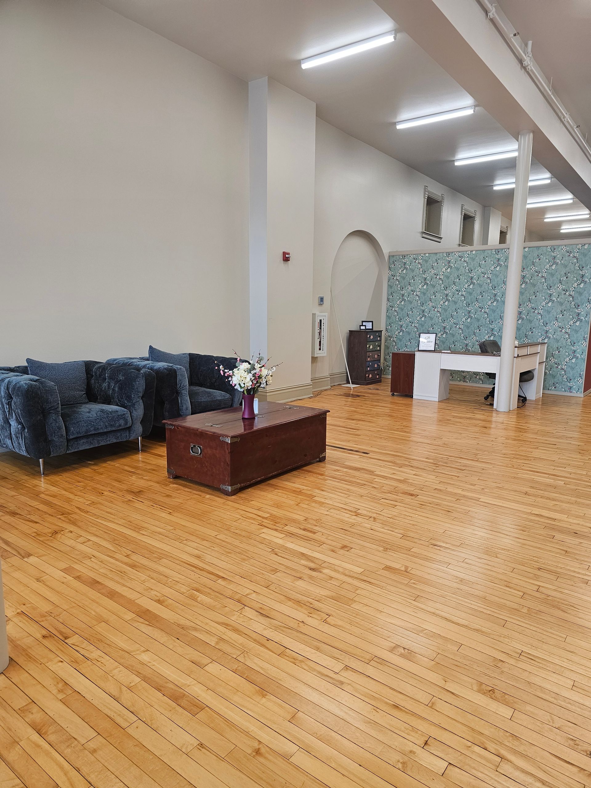 Spacious office with hardwood floors, blue sofa, dark wooden coffee table, white desk, and light-colored walls.