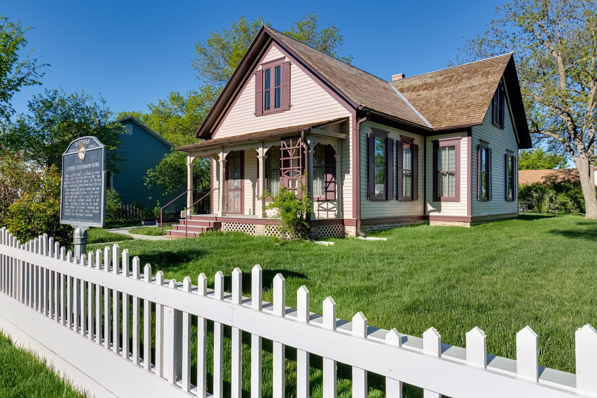 White picket fence in front of a pink Victorian house with a porch and green lawn under a blue sky